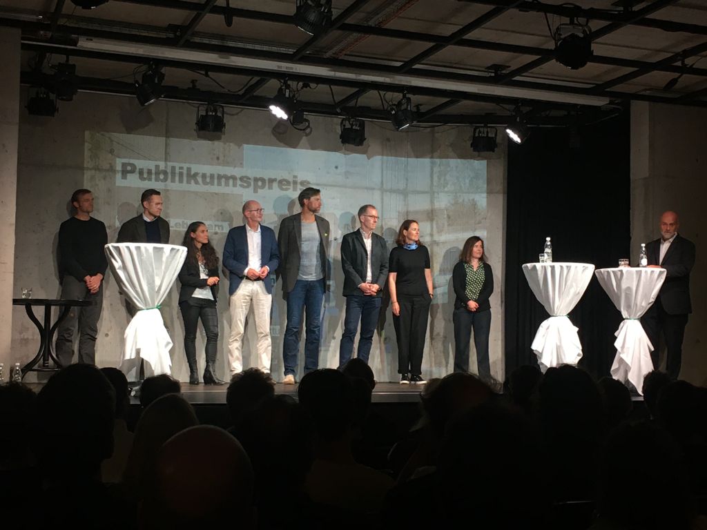 Acht Personen auf einem Podium wird von einer Person ein Preis verleihen