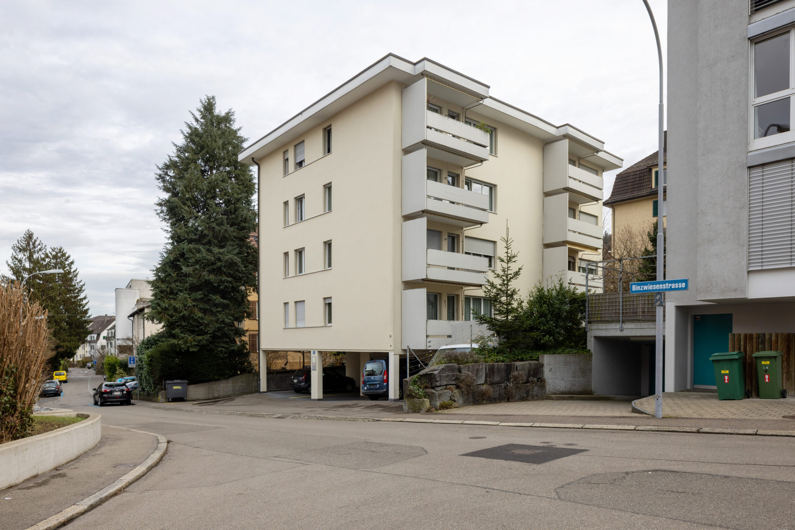 Strassenansicht der Binzwiesenstrasse 12. Die Westseite des Hauses mit den zwei Balkonreihen ist zu sehen. Hinter dem Haus ist ein grosser Baum.