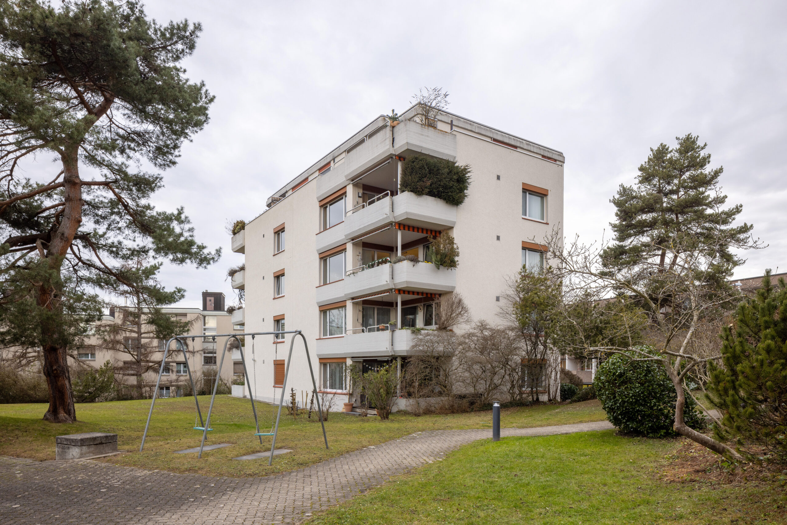 Wiesliacher 45 hofseitig die Ecke mit bewachsenen Balkonen abgebildet. Grünflächen, Bäume, ein kleiner Weg und eine Schaukel umgeben das Haus.