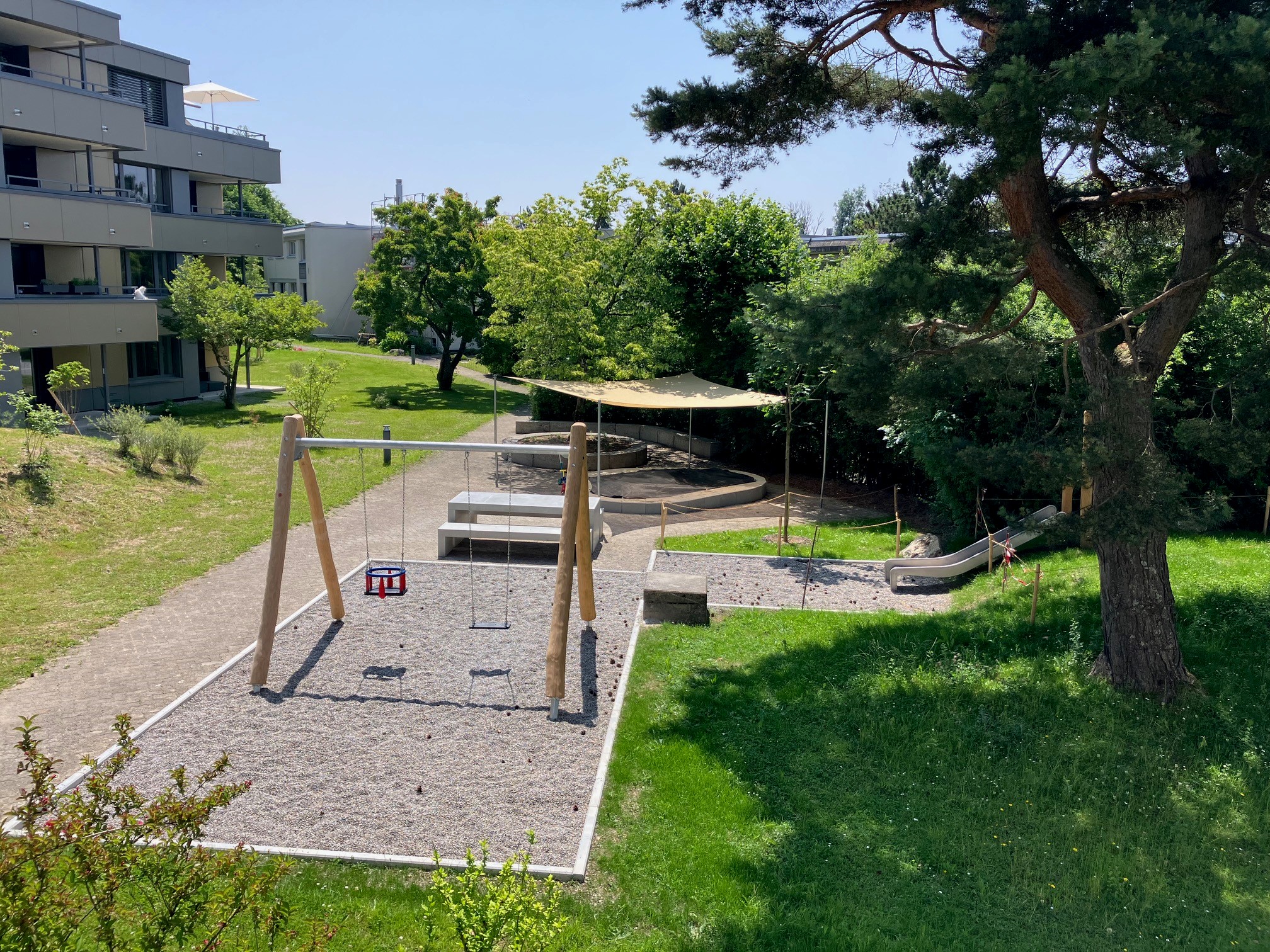 Spielplatz Wiesliacher 45 mit Schaukel, Rasen, Rutsche, Steintisch und - bank, und überdecktem Sandkasten.